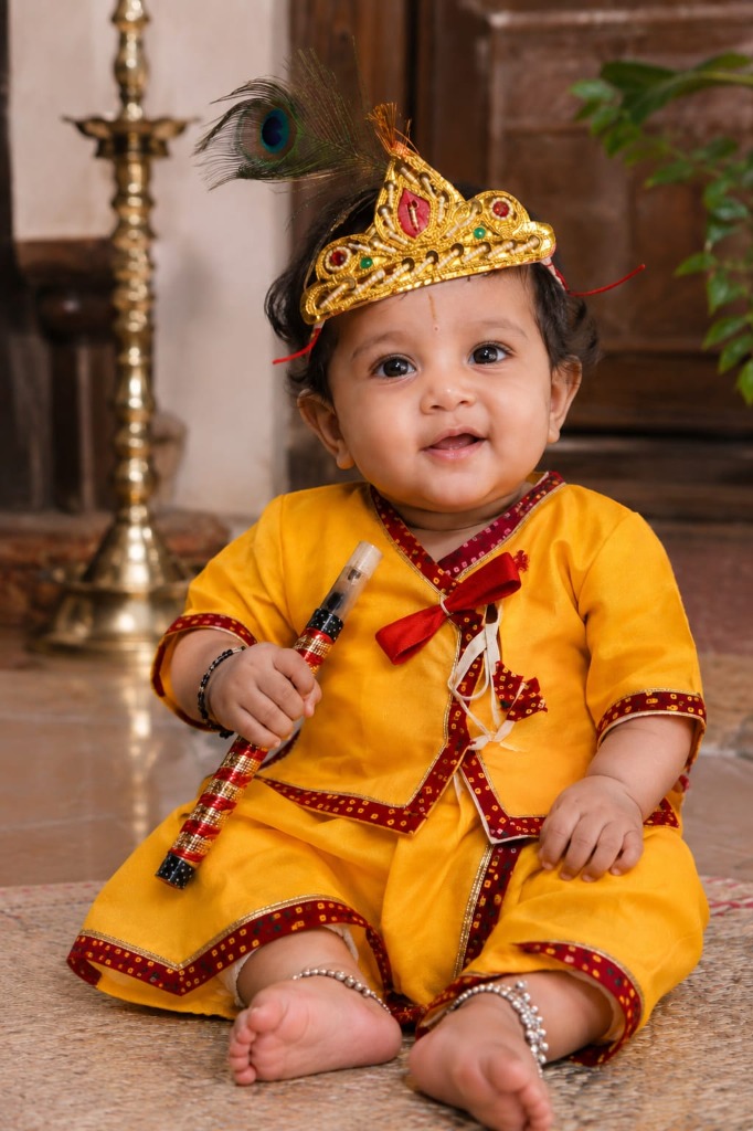 Baby boy sitting comfortably in a soft cotton yellow Krishna dress with mukut and flute from Mummy Daddy Readymades.