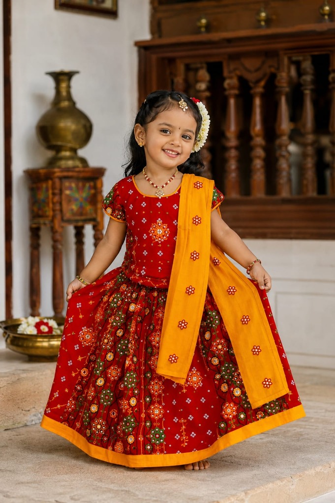 Young girl smiling in a red and green pavadai style Radha dress with a pinned yellow dupatta from Mummy Daddy Readymades.